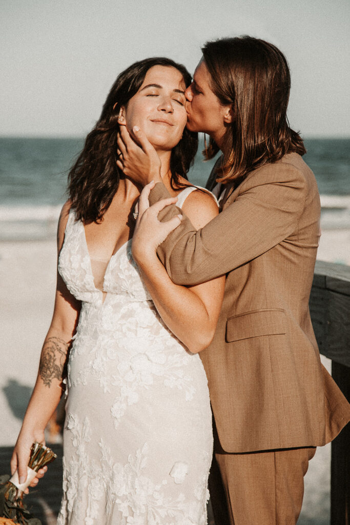 Bride kissing other bride on the check in direct sunlight wit the beach behind them during their Florida elopement