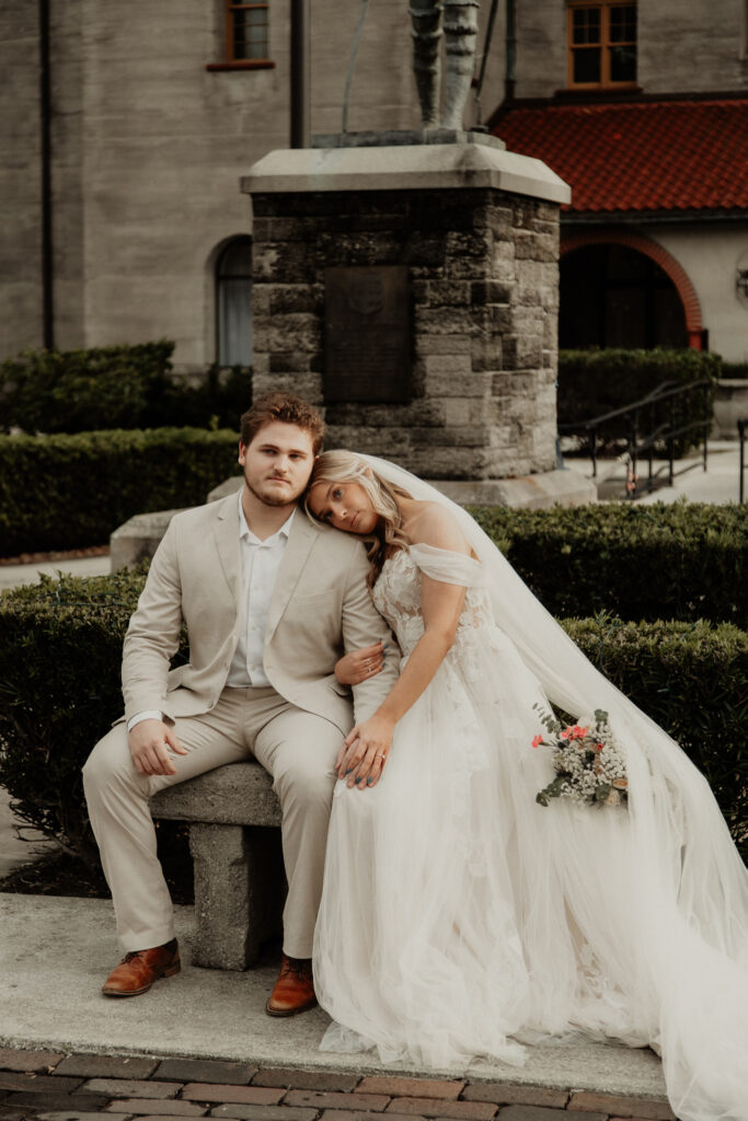 bride and groom are sitting on a bench in front of the lightner museum. bride is resting her head on the grooms shoulder with her flowers next to her side