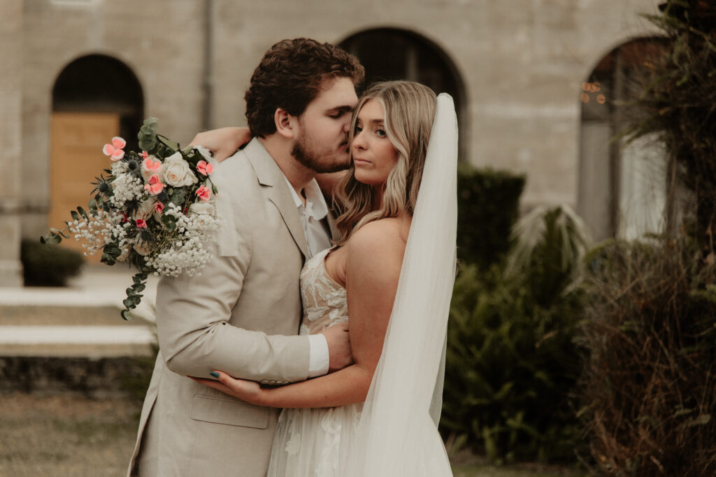 Groom is kissing the bride on the cheek while the bride looks off in the distance.