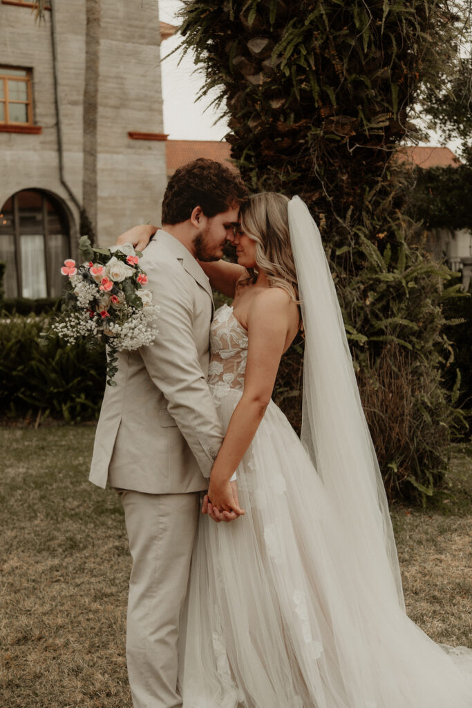 A bride and groom share an intimate moment, foreheads touching and hands intertwined. The bride holds a bouquet with one hand while resting her other hand on the groom's shoulder