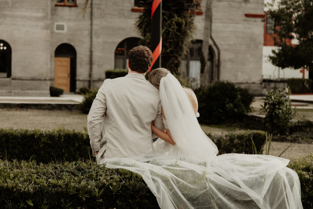 bride and groom are leading up against the bushes. Bride is leaning her head on his shoulder